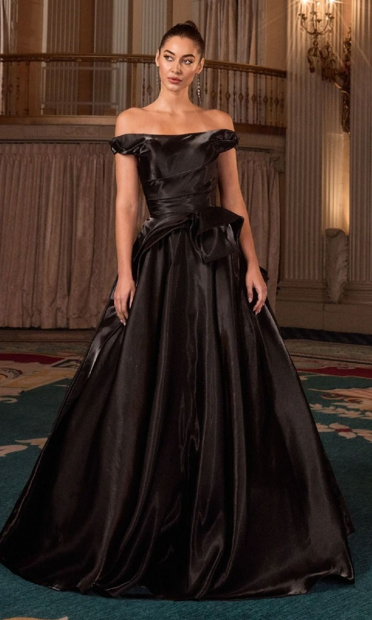 A woman stands indoors wearing the Ladivine J885, a long prom dress featuring an off-the-shoulder satin bodice, full A-line skirt, and a bow detail at the waist.